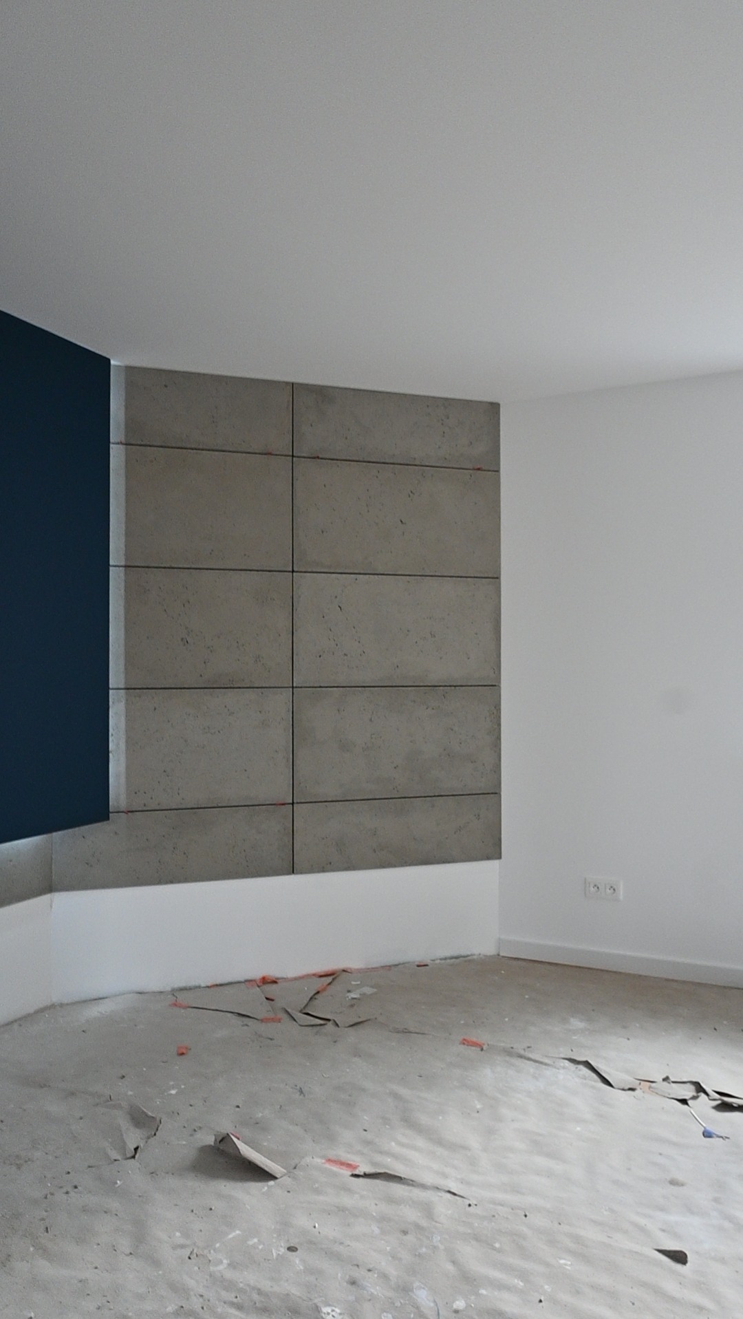 Ściana wykończona betonowymi panelami w surowym stylu, z fragmentem pomalowanym na granatowo. Podłoga zabezpieczona folią malarską. Widoczne gniazdka elektryczne.