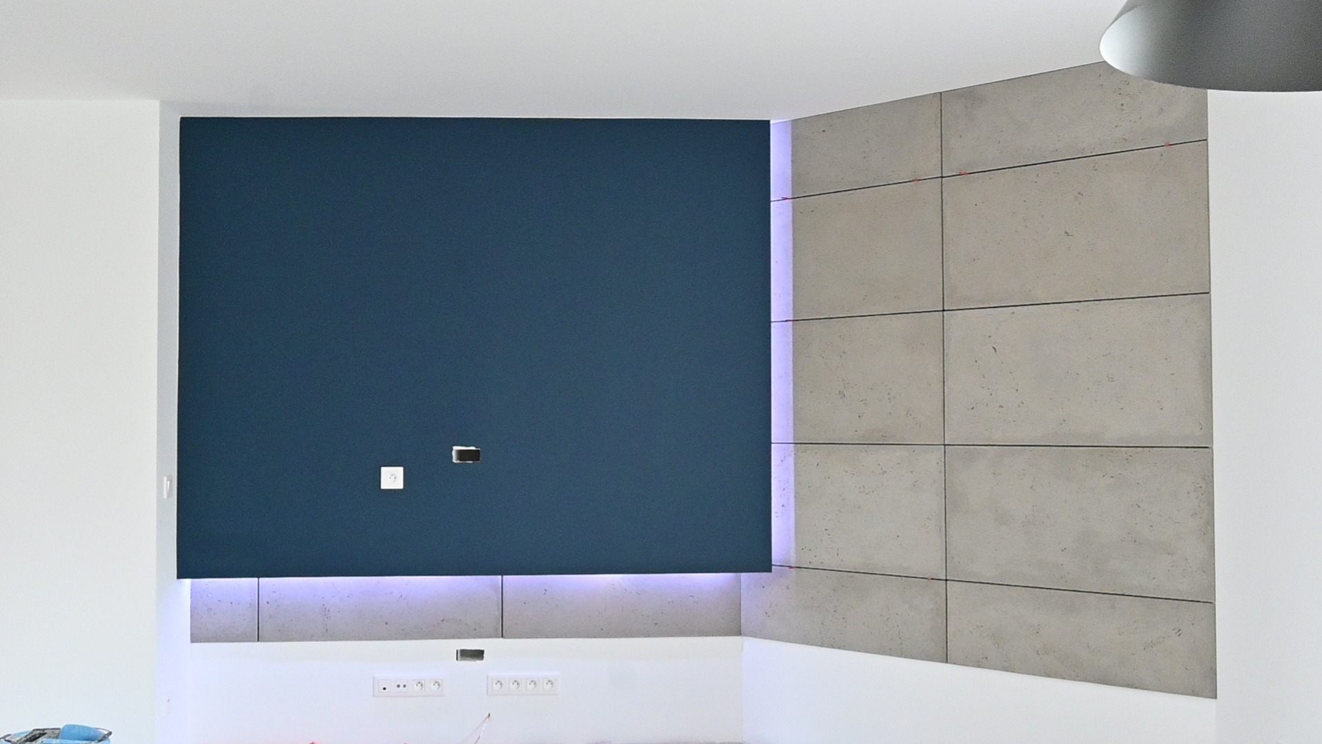 Niebieska ściana z gładzią i betonowe płyty dekoracyjne z podświetleniem LED w nowoczesnym wnętrzu. Widoczne gniazdka elektryczne i puszki instalacyjne.