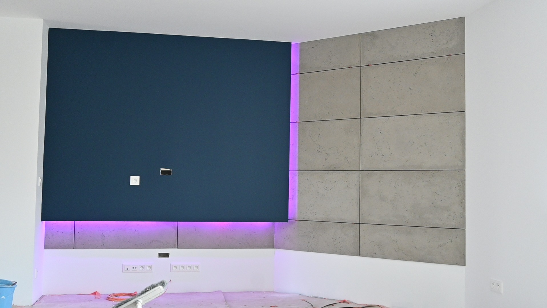 Narożnik pokoju z panelami dekoracyjnymi: ciemnoniebieski materiał i betonowe płyty, podświetlone fioletowym światłem LED. Widoczne gniazdka elektryczne i narzędzia.