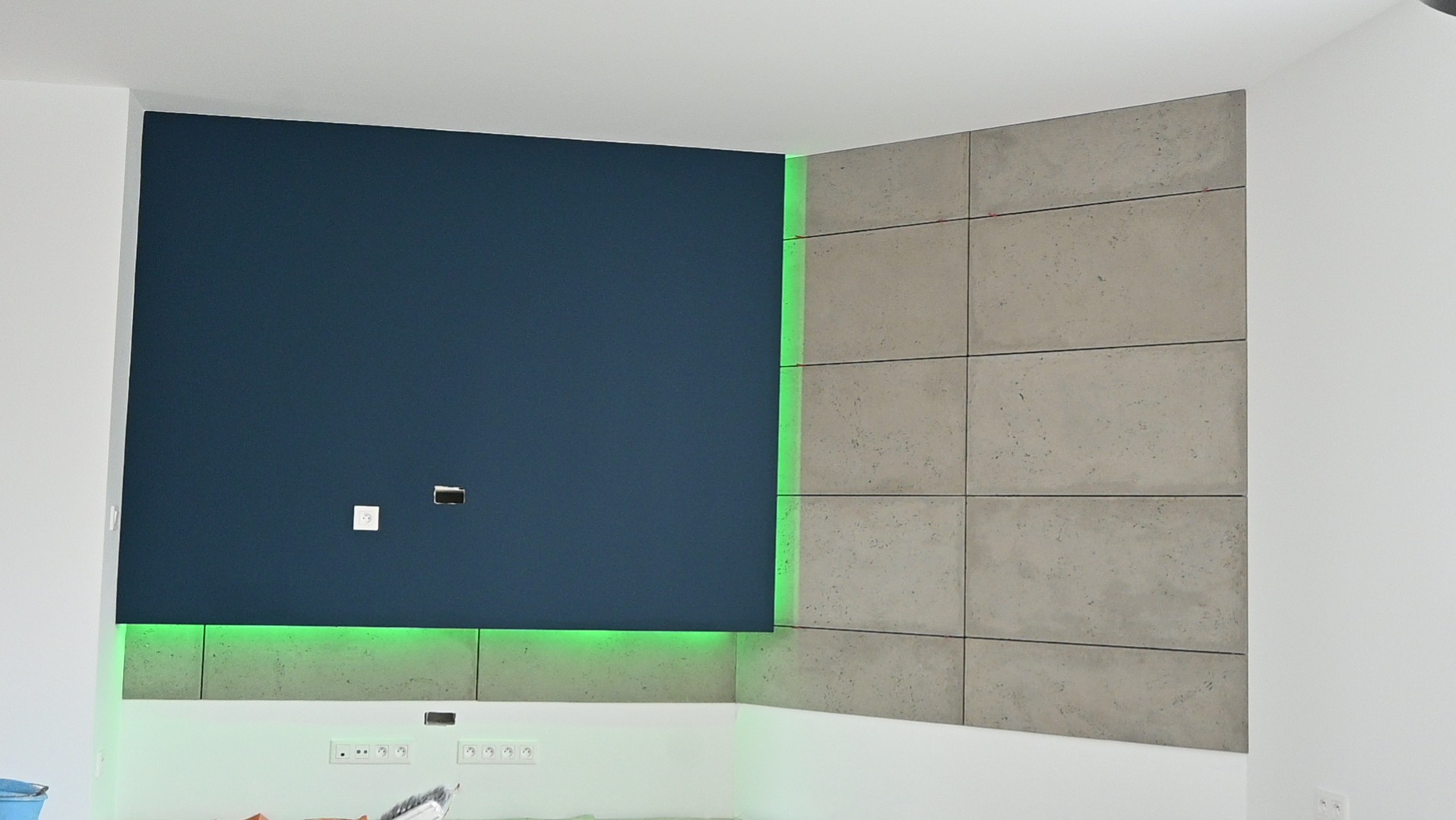 Niebieska ściana z podświetleniem LED i szare panele dekoracyjne tworzą nowoczesny design wnętrza. Widoczne gniazdka elektryczne i otwory na kable.