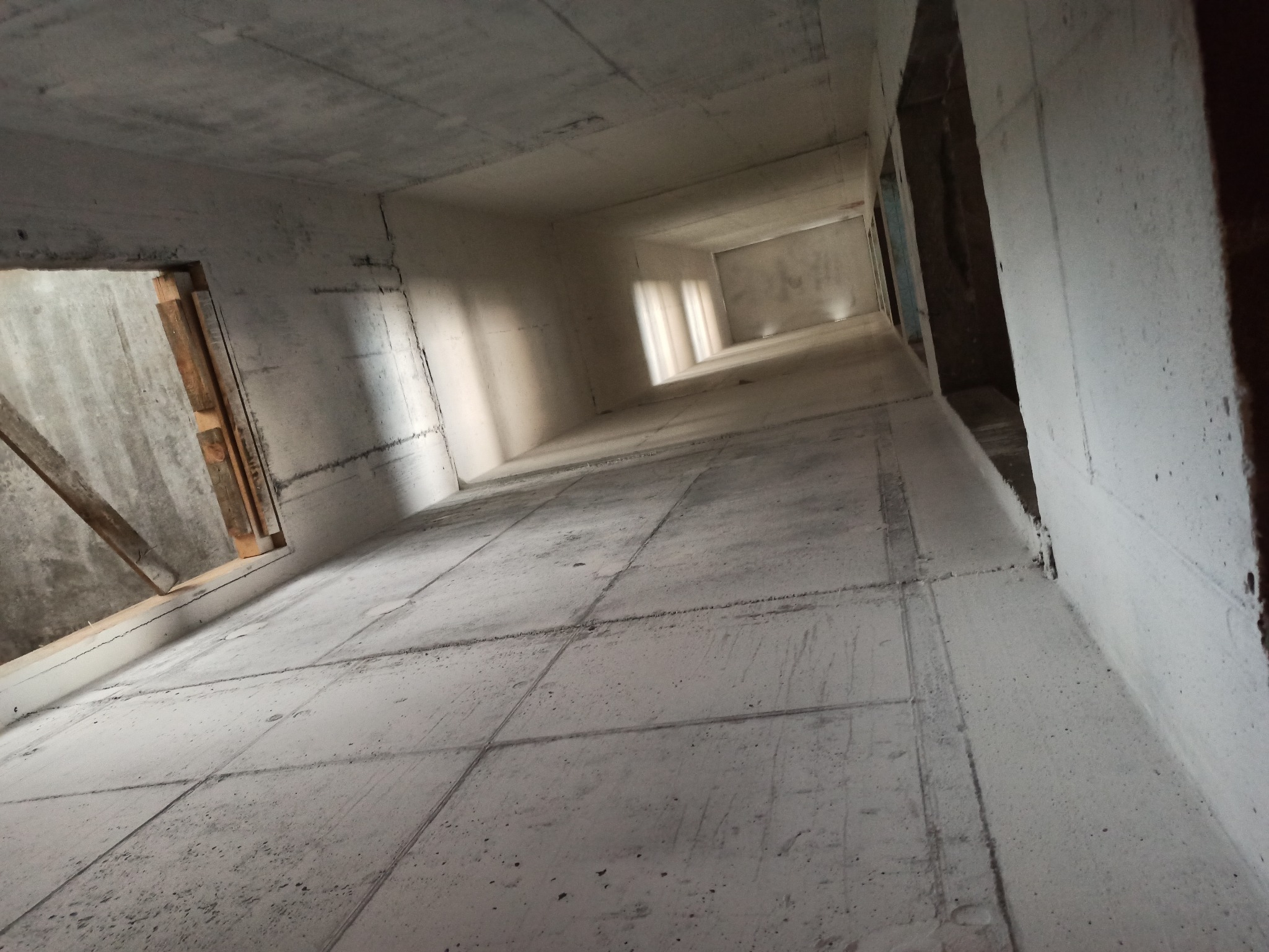 Surowe wnętrze w trakcie budowy z widocznymi betonowymi ścianami, podłogą z płyt i surowym oknem z drewnianymi elementami, wpadające światło dzienne.