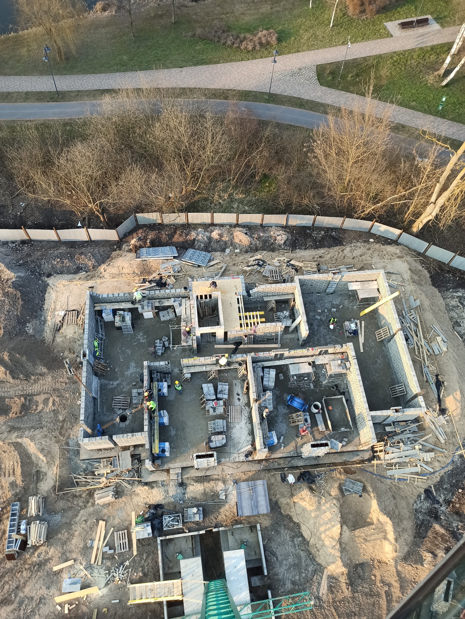 Widok z góry na plac budowy domu z częściowo wzniesionymi ścianami z bloczków, widoczni robotnicy w kaskach, materiały budowlane i otoczenie z drzewami oraz ścieżką.