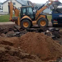 Żółta koparko-ładowarka CAT załadowuje gruz i ziemię na niebieską wywrotkę z widoczną tablicą rejestracyjną GKA 21JT, w tle budynki.