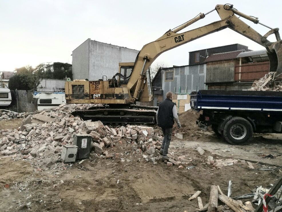 Żółta koparka CAT załadowuje gruz do niebieskiej ciężarówki; w tle białe przyczepy kempingowe i budynki, a na pierwszym planie mężczyzna w szarej kamizelce.