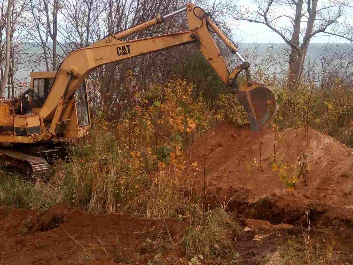 Żółta koparka CAT w trakcie pracy na zalesionym terenie, z widocznym wykopem i stertą ziemi, tło stanowią drzewa i odległy zbiornik wodny.
