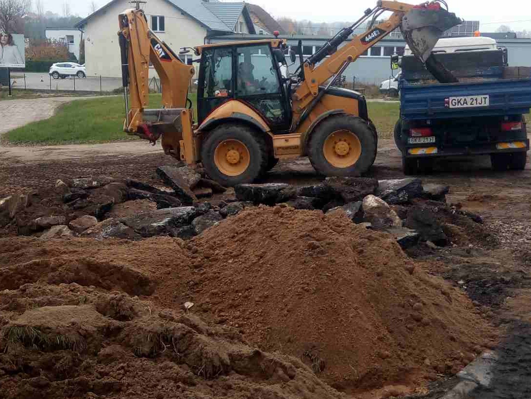 Żółta koparko-ładowarka CAT załadowuje gruz i ziemię na niebieską wywrotkę z widoczną tablicą rejestracyjną GKA 21JT, w tle budynki.