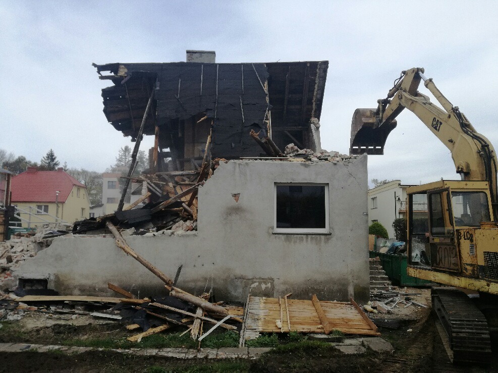 Żółta koparka CAT podczas rozbiórki częściowo zawalonego, dwukondygnacyjnego budynku z widocznymi fragmentami spalonych elementów konstrukcyjnych. W tle zabudowa mieszkalna i pochmurne niebo.