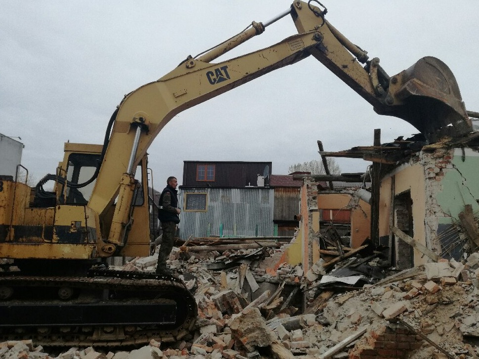 Żółta koparka CAT w trakcie rozbiórki budynku, operator w kamizelce stoi na gąsienicach, w tle zabudowa miejska i pochmurne niebo.