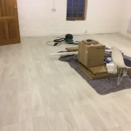 Jasne panele podłogowe ułożone w pustym pokoju z białymi ścianami, widoczne kartony z panelami, odkurzacz i krzesło w tle.