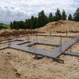 P.H.U ERGONOMECO BAU Krzysztof Wdowiak - Zalane betonem szalunki fundamentowe z wystającymi prętami zbrojeniowymi, otoczone piaskiem, z hałdą piasku w tle i zielonym krajobrazem w oddali, w słoneczny dzień.