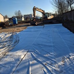 Trans-Kop Bartłomiej Kamiński - Żółta koparka Liebherr na placu budowy, w tle widoczny pracownik w odblaskowej kamizelce i przenośna toaleta, a na pierwszym planie rozłożona biała geowłóknina z odciskami butów.