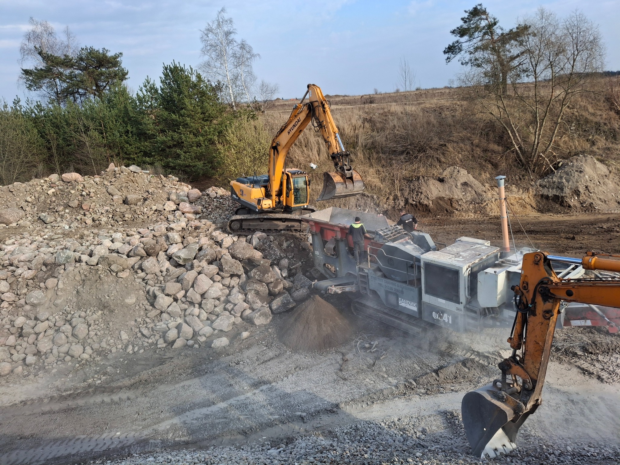 Żółta koparka Hyundai ładuje kamienie do kruszarki Sandvik na placu budowy, w tle drzewa i niebo