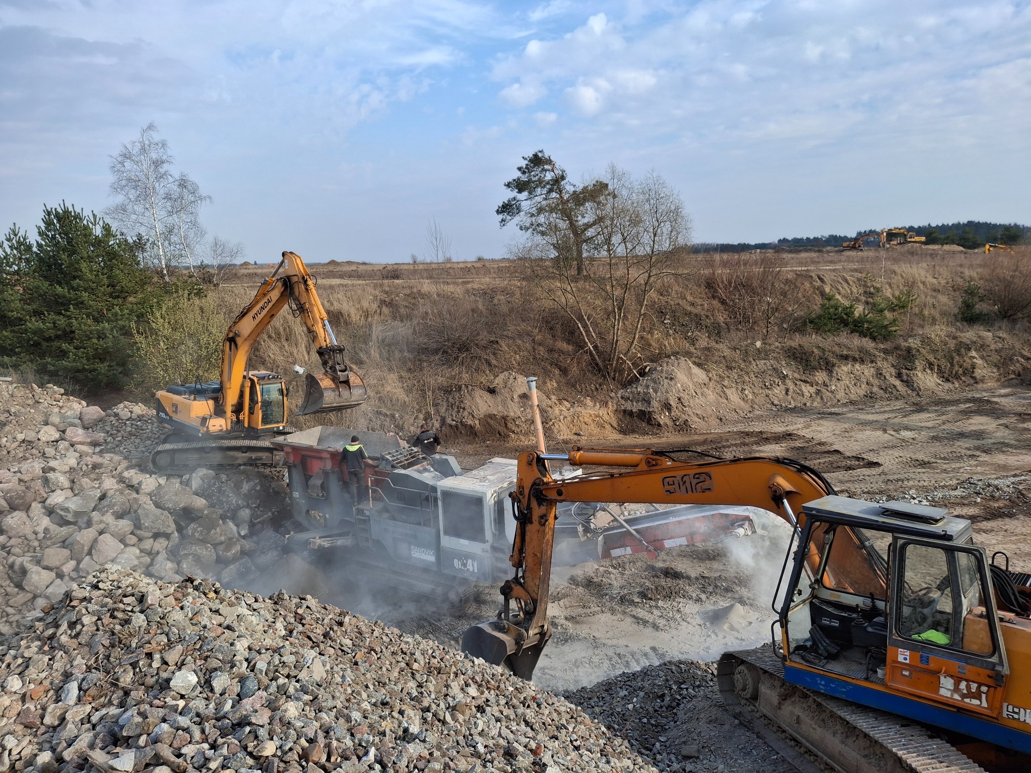 Dwie pomarańczowe koparki Hyundai i przesiewacz mobilny Sandvik na placu budowy; w tle widoczne drzewa i błękitne niebo z chmurami.