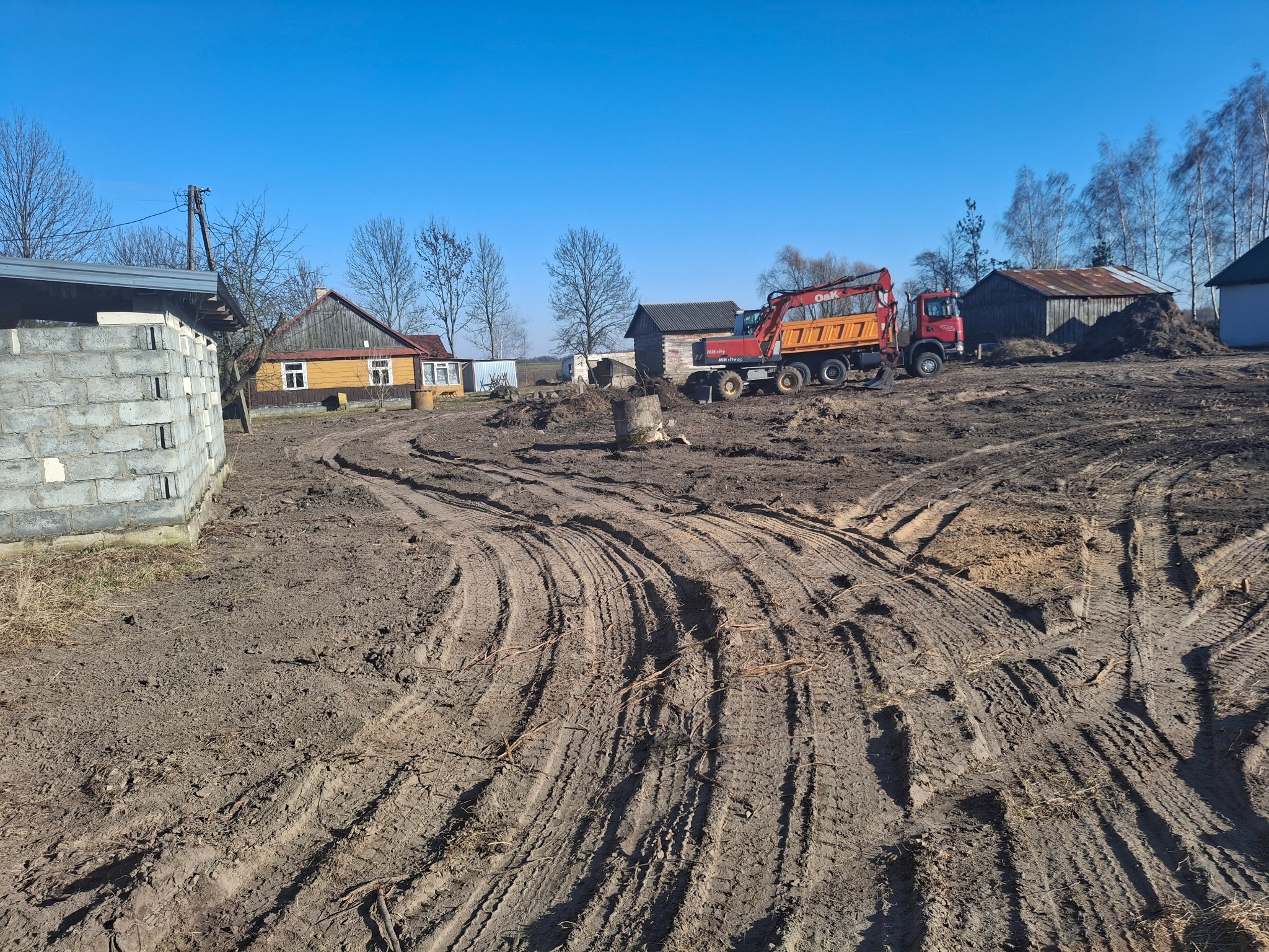Ciężarówka z wywrotką i koparka kołowa na placu budowy z widocznymi śladami opon w błotnistym terenie, w tle budynki gospodarcze i domy mieszkalne w słoneczny dzień.