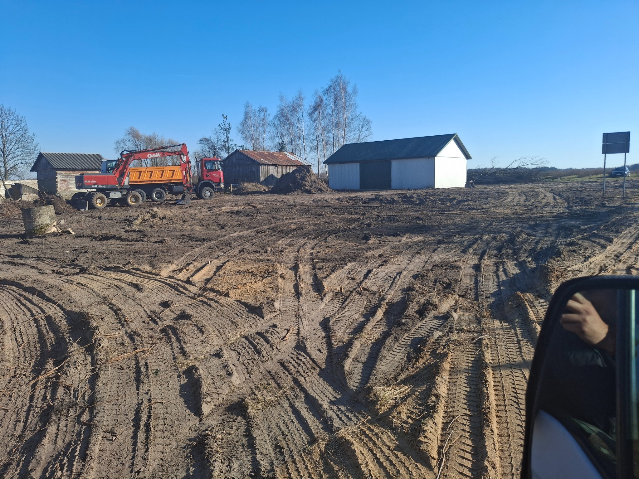 Czerwona koparko-ładowarka i ciężarówka na placu budowy, widoczne ślady opon na ziemi, w tle budynki gospodarcze i drzewa pod błękitnym niebem.