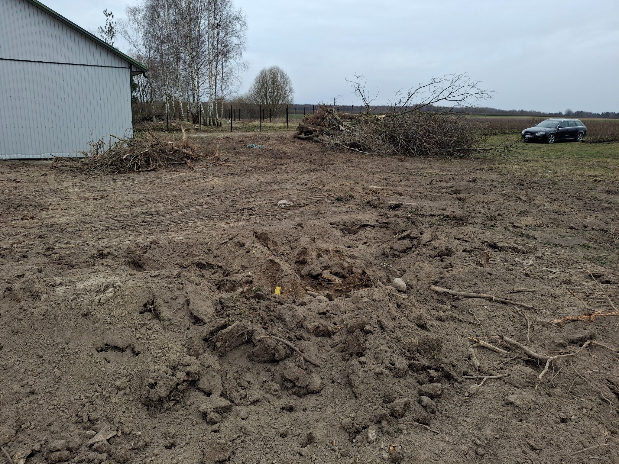 Przygotowany teren pod budowę, z usuniętymi drzewami i wykopanym dołem, sterta gałęzi i ziemi, w tle budynek gospodarczy i samochód osobowy.