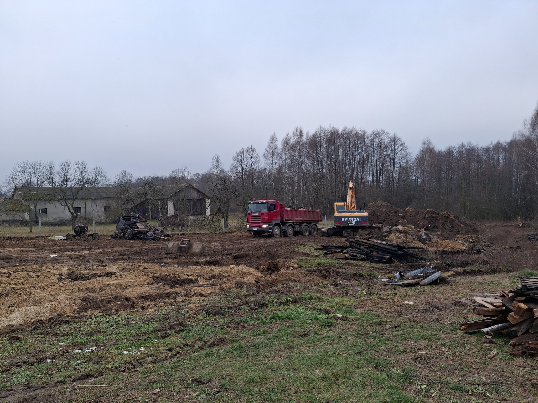 Czerwona ciężarówka Scania i koparka Hyundai na placu budowy z rozkopaną ziemią, w tle budynki gospodarcze i drzewa bez liści pod pochmurnym niebem.