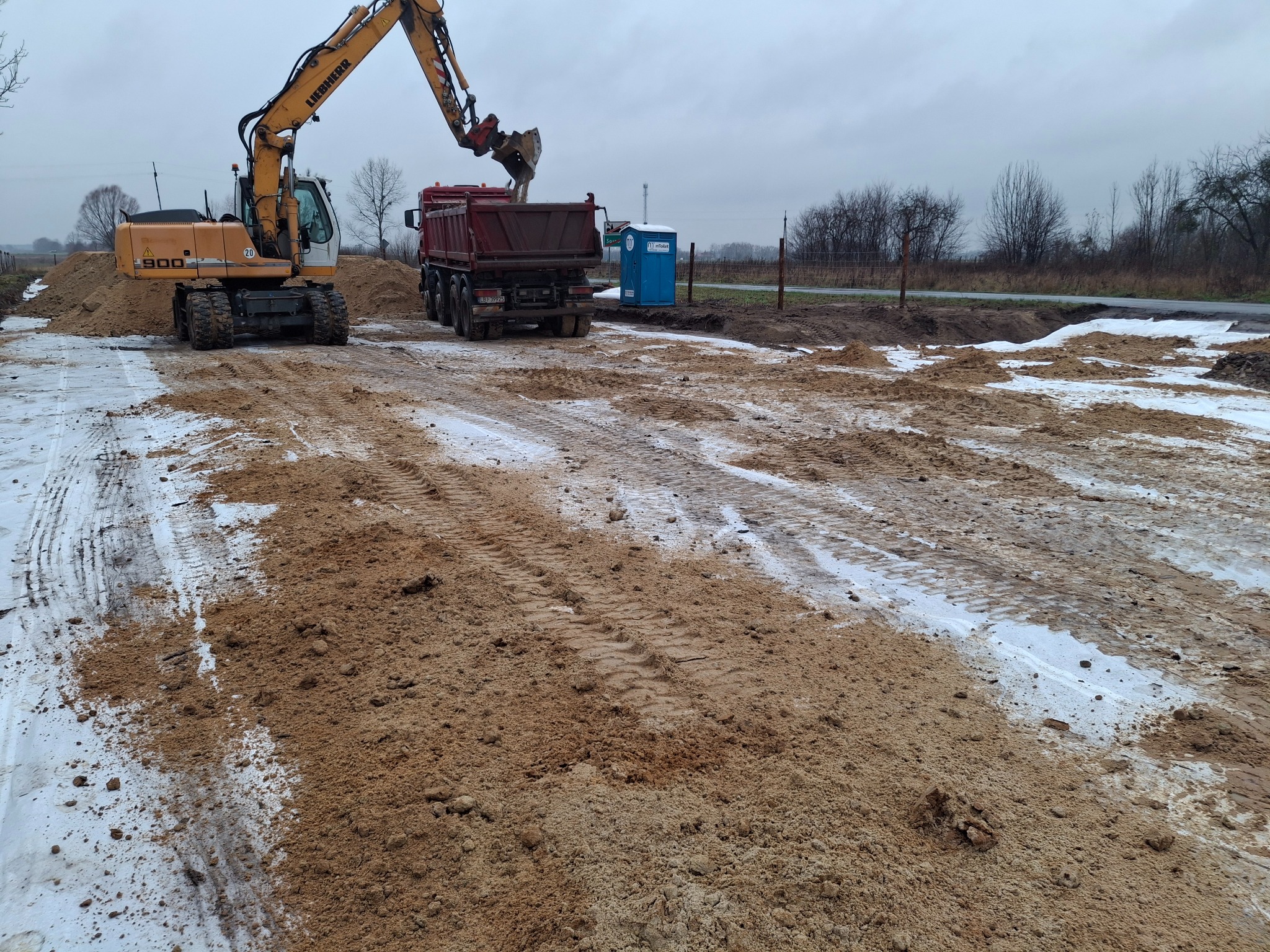 Żółta koparka Liebherr 900 ładuje piasek do czerwonej wywrotki na placu budowy z widoczną przenośną toaletą w tle.