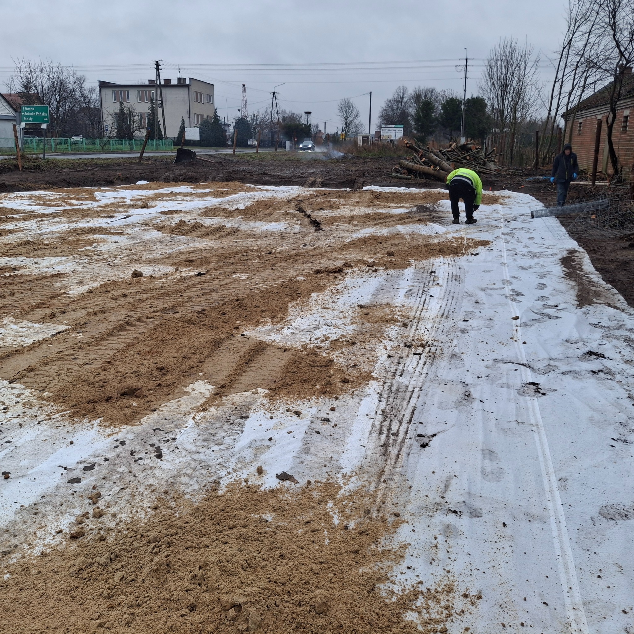 Przygotowanie terenu pod budowę: rozłożona geowłóknina częściowo zasypana piaskiem, widoczne ślady opon, pracownik w odblaskowej kamizelce i drugi pracownik z rolką siatki ogrodzeniowej w tle...
