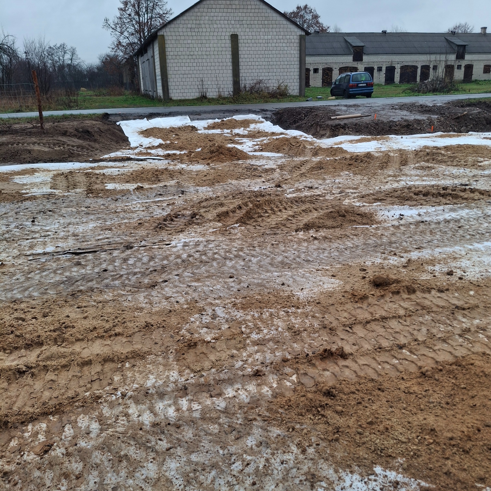 Przygotowany teren pod budowę, z widocznymi śladami opon ciężkiego sprzętu i białą geowłókniną, w tle zabudowania gospodarcze.