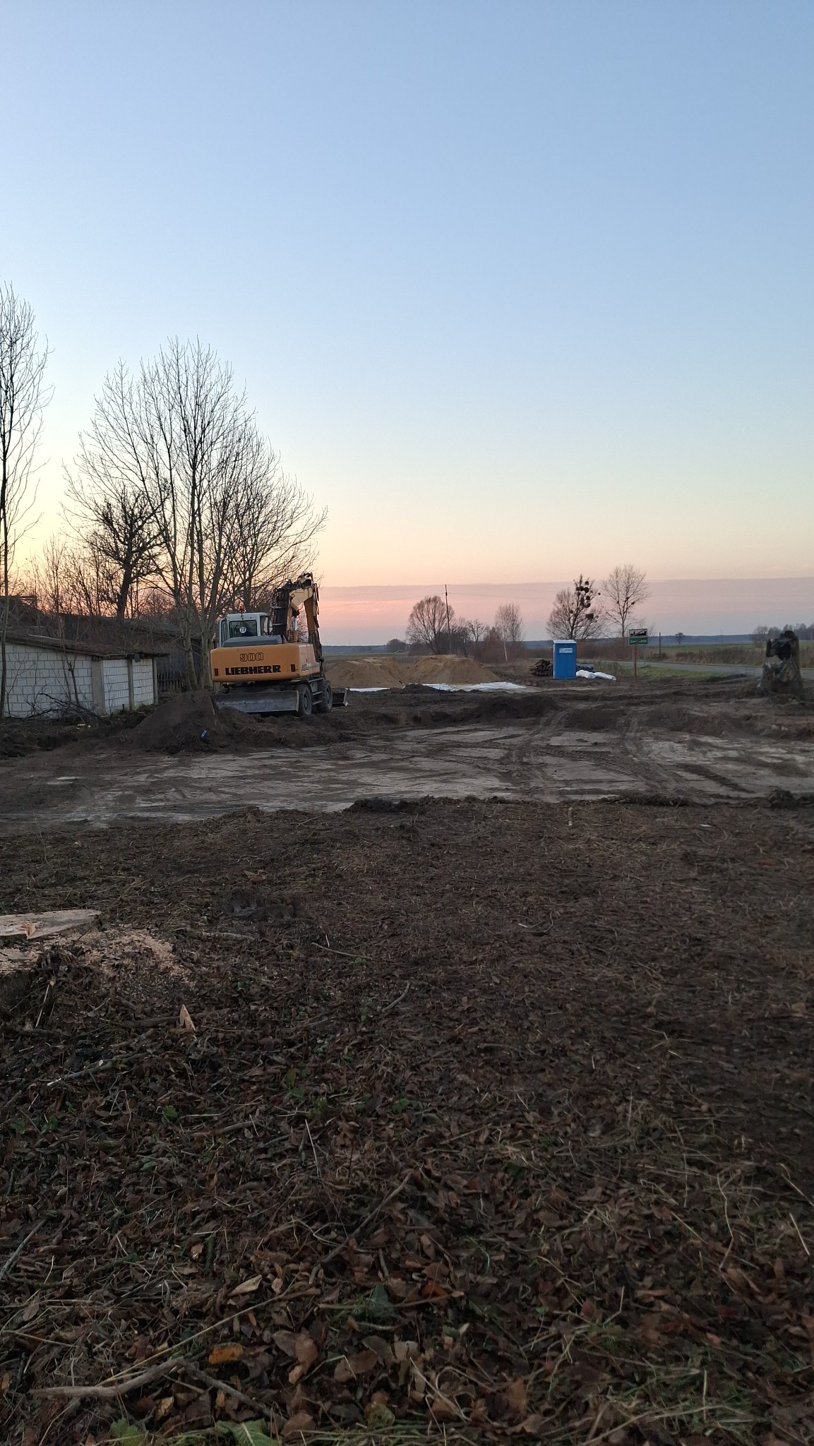 Żółta koparka Liebherr 900 na placu budowy o zmierzchu, widoczne ślady opon na ziemi, w tle budynek gospodarczy z białej cegły i błękitna toaleta przenośna.