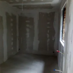 Pokój w trakcie remontu: ściany i sufit pokryte płytami gipsowo-kartonowymi, widoczne szpachlowane łączenia, wystające przewody elektryczne, okno w trakcie montażu.