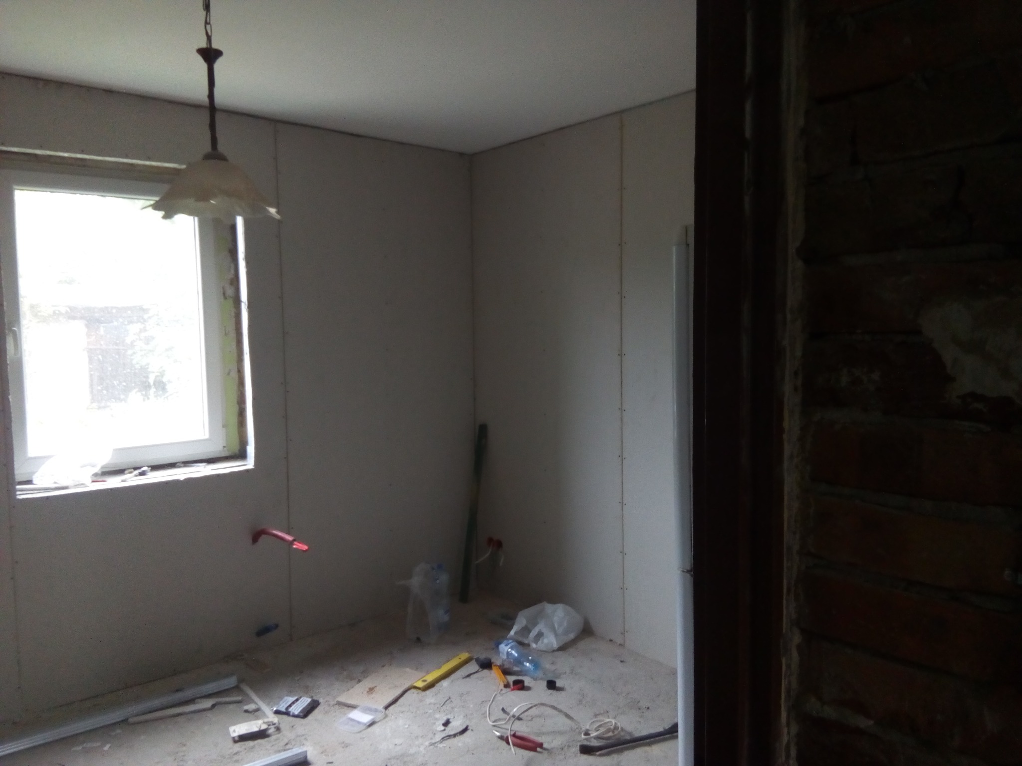 Wnętrze w trakcie montażu ścianek działowych z płyt gipsowo-kartonowych, widoczne narzędzia i materiały budowlane na podłodze, stara lampa wisząca i fragment muru z cegły w tle.