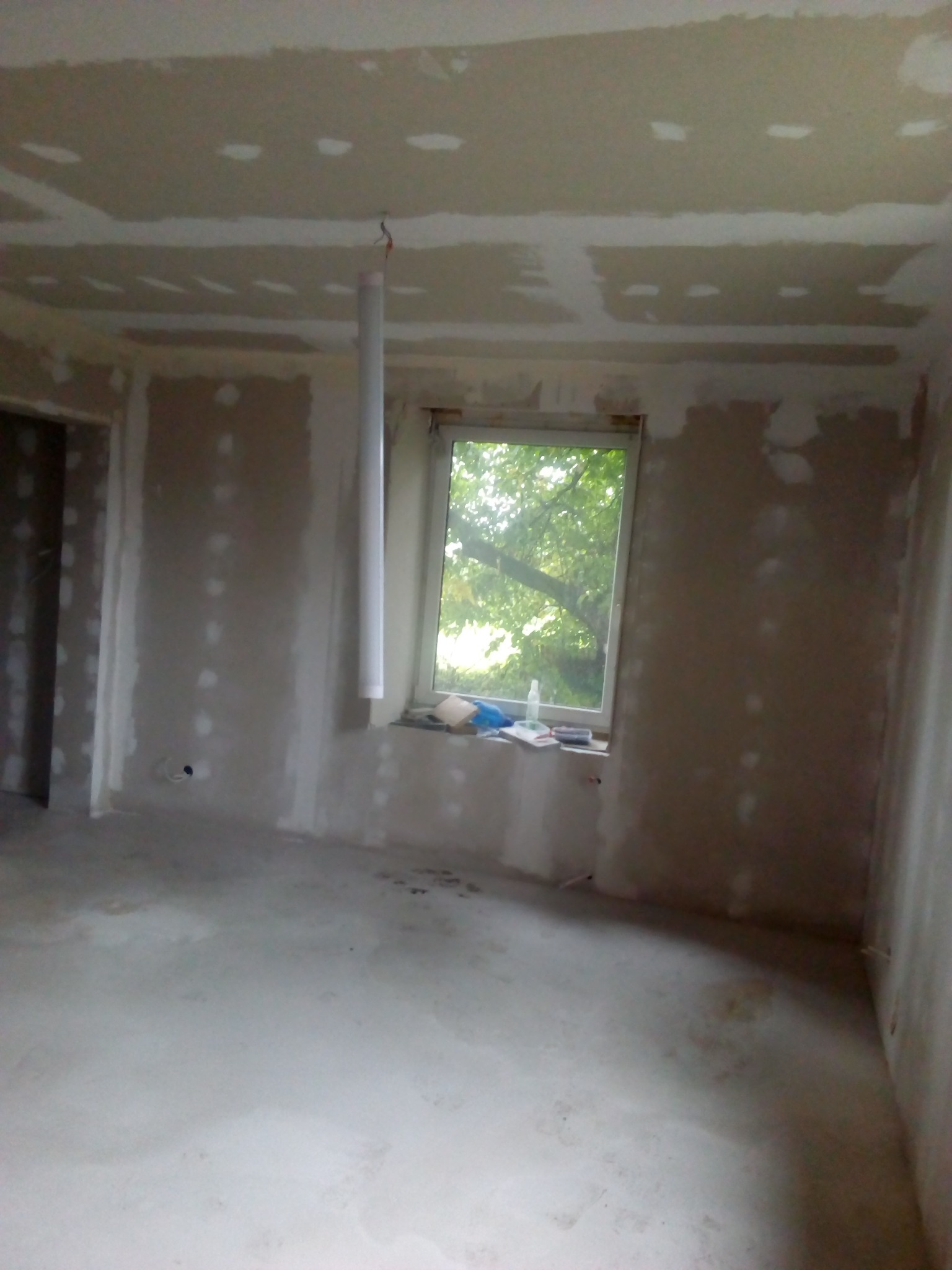 Pokój w trakcie prac wykończeniowych: ściany i sufit pokryte gładzią gipsową, widoczne łączenia płyt gipsowo-kartonowych, rura wentylacyjna zwisająca z sufitu, okno z widokiem na zieleń.