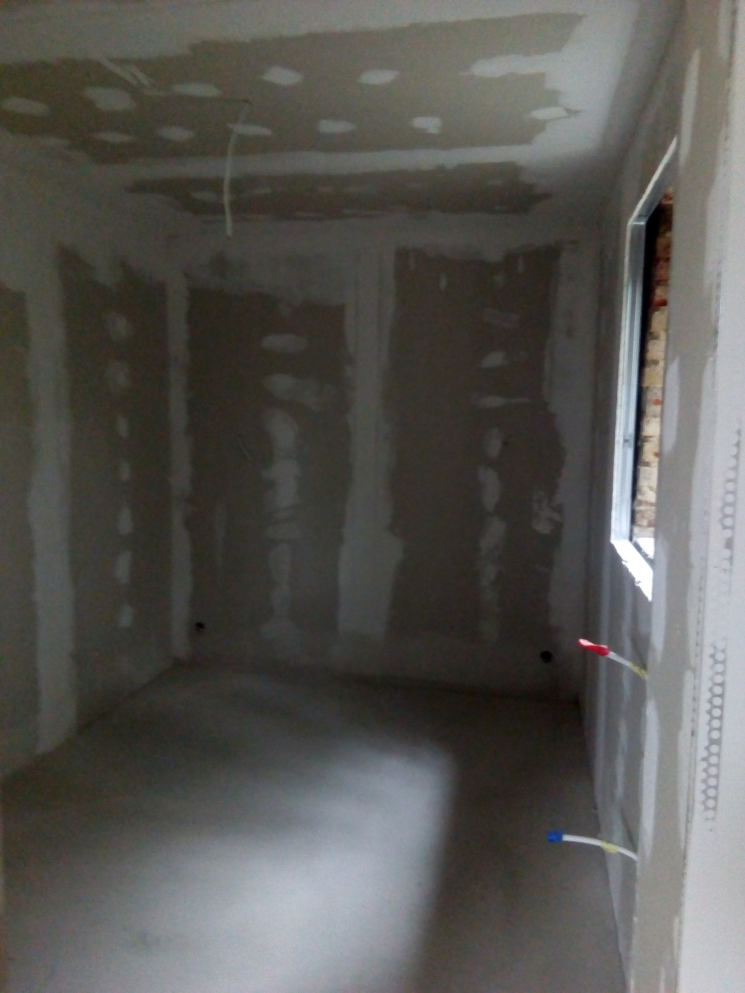 Pokój w trakcie remontu: ściany i sufit pokryte płytami gipsowo-kartonowymi, widoczne szpachlowane łączenia, wystające przewody elektryczne, okno w trakcie montażu.