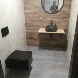 Magdalena Stępień - Nowoczesna łazienka z czarną wiszącą toaletą i umywalką nablatową, drewnopodobne płytki na ścianie za umywalką, szare płytki na podłodze, w lustrze odbicie osoby robiącej zdjęcie.