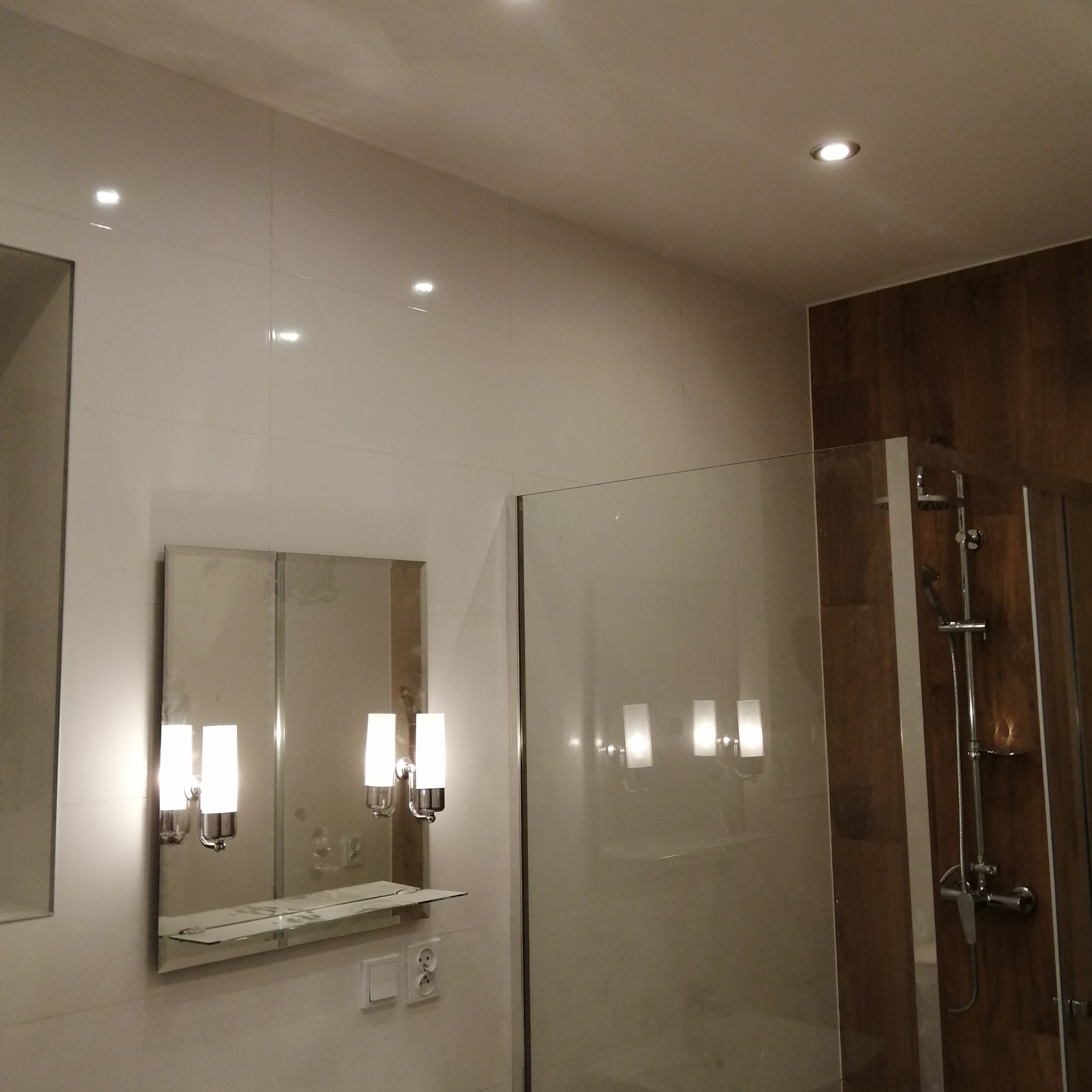 Wykończona łazienka z jasnymi, błyszczącymi płytkami ściennymi, szklaną kabiną prysznicową z chromowaną armaturą i lustrem z oświetleniem kinkietowym.