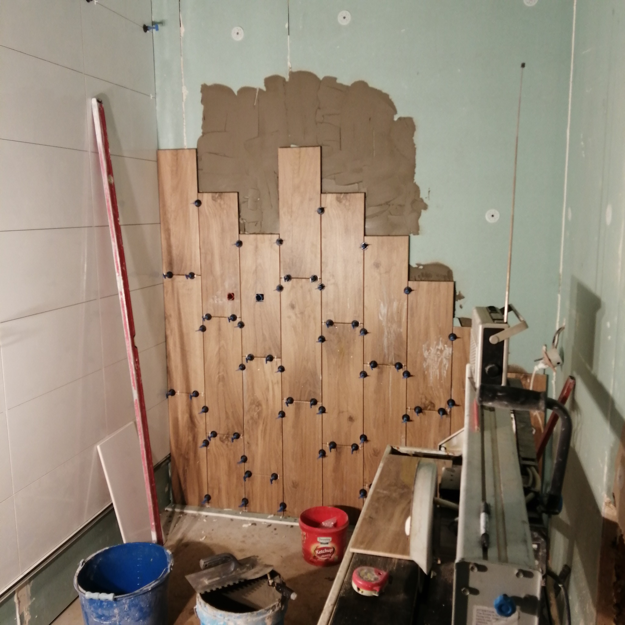 Układanie drewnopodobnych płytek na ścianie z użyciem krzyżyków dystansowych, widoczna zaprawa, poziomica, wiaderka i przecinarka do płytek.