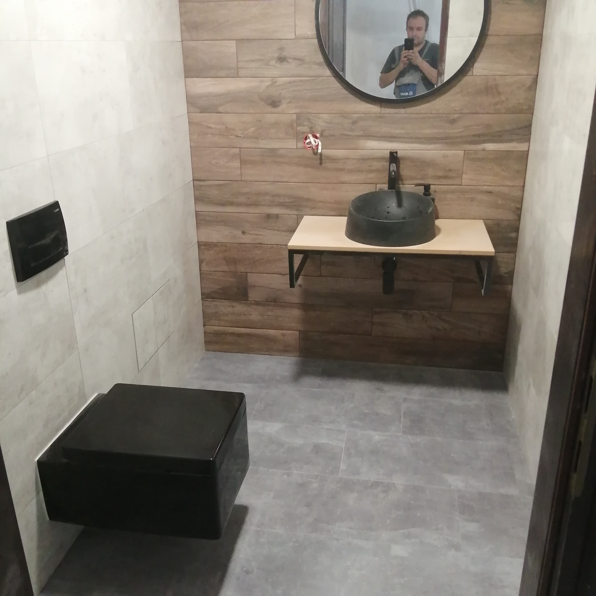 Nowoczesna łazienka z czarną wiszącą toaletą i umywalką nablatową, drewnopodobne płytki na ścianie za umywalką, szare płytki na podłodze, w lustrze odbicie osoby robiącej zdjęcie.