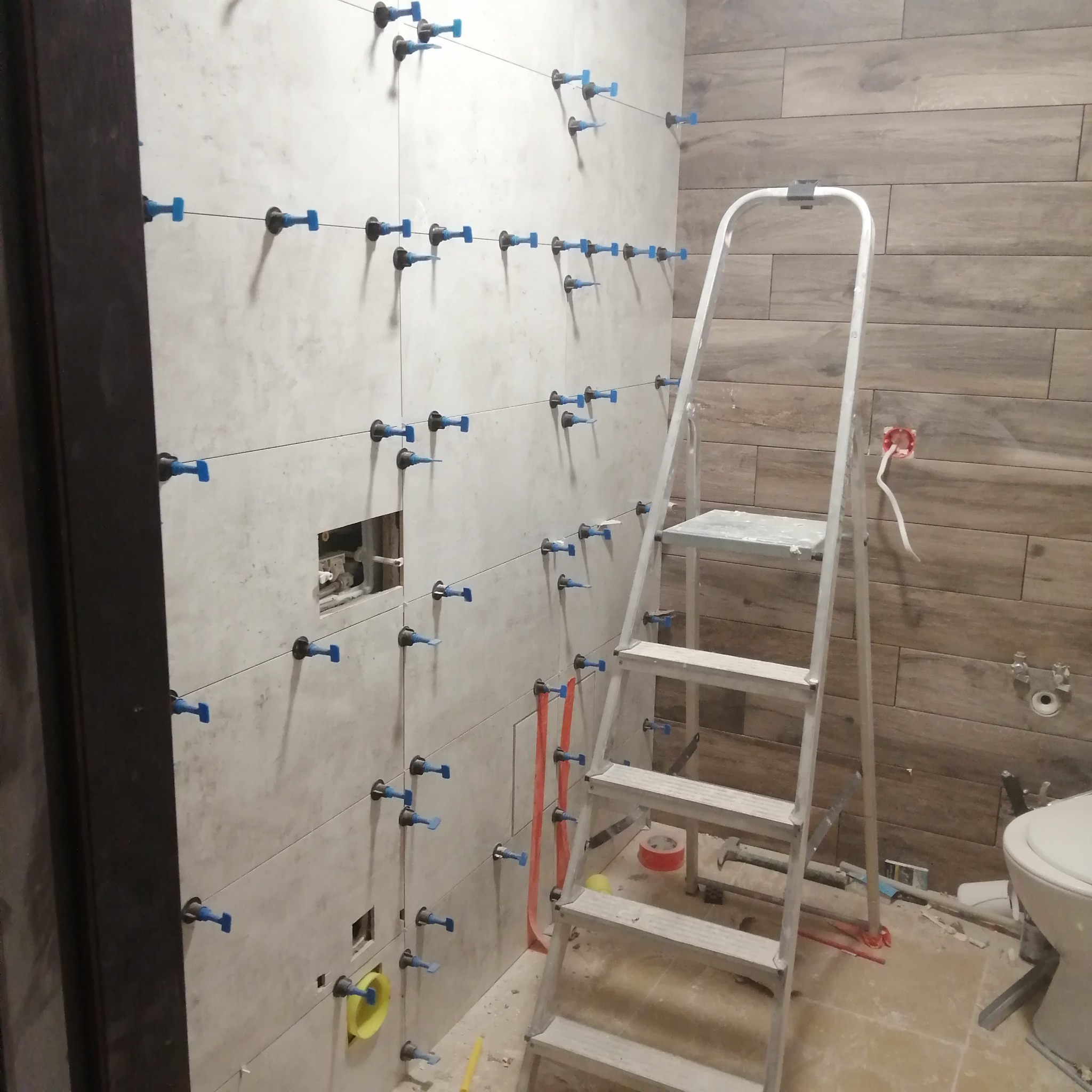 Świeżo ułożone, jasne płytki na ścianie łazienki, zabezpieczone niebieskimi klipsami poziomującymi, widoczna aluminiowa drabina, fragment toalety i instalacji elektrycznej.