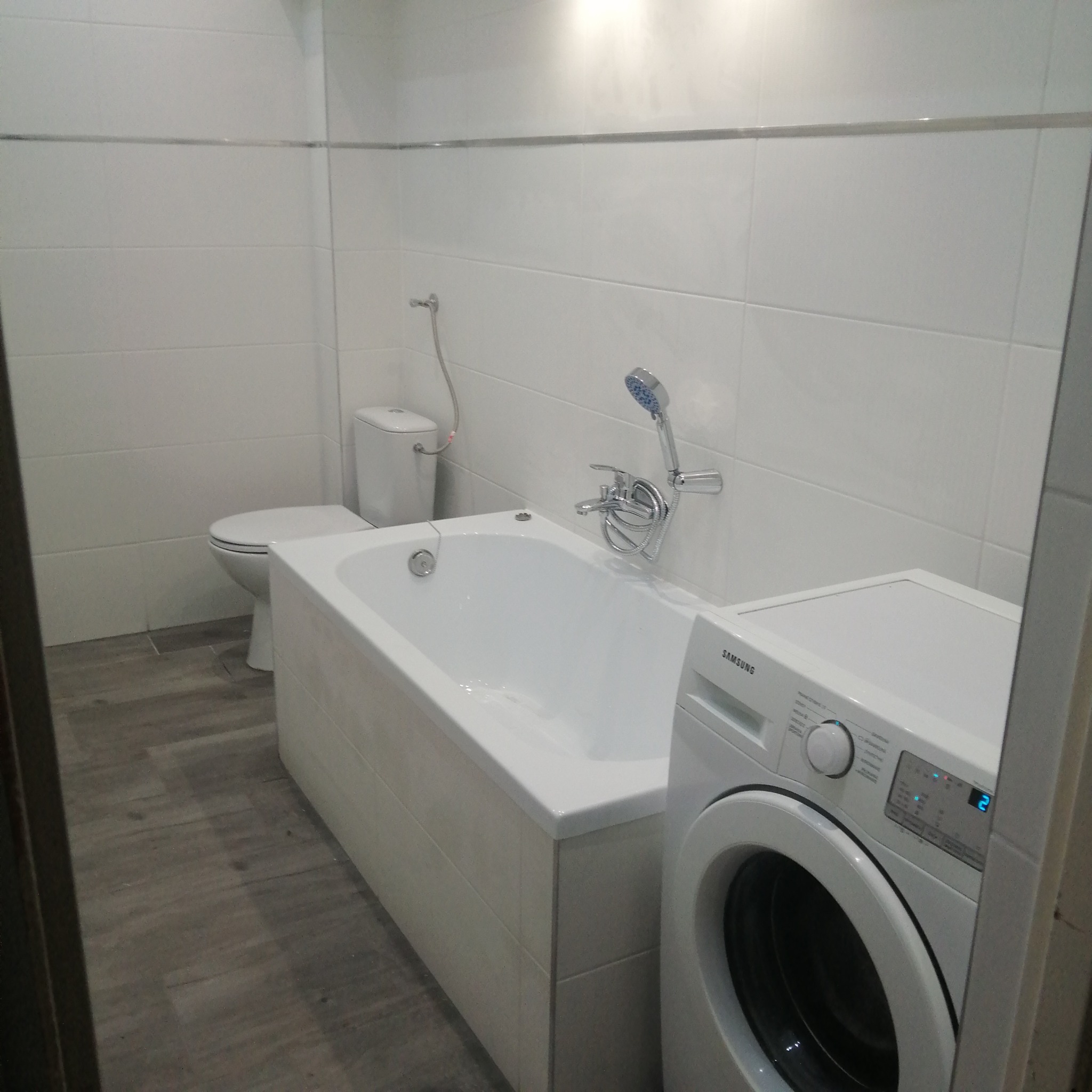 Wykończona łazienka z białą armaturą, płytkami na ścianach i podłodze imitującymi drewno, pralka Samsung widoczna z prawej strony.