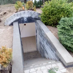 Wejście do piwnicy ziemianki z betonowych bloków, częściowo wykończone płytkami, otoczone roślinnością.