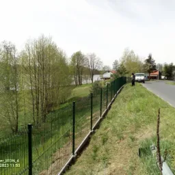 Nowo zainstalowane, zielone ogrodzenie panelowe z podmurówką betonową, oddzielające posesję od drogi asfaltowej i jeziora w tle; widoczny samochód dostawczy i osoba stojąca obok.