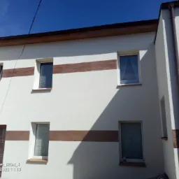 Fragment fasady budynku z białym tynkiem i poziomymi pasami imitującymi drewno, widoczne okna z białymi ramami oraz brązowe rynny.