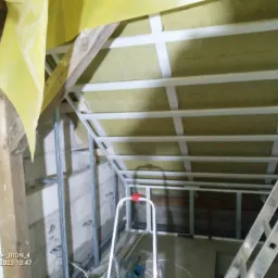 Poddasze w trakcie ocieplania wełną mineralną, widoczne metalowe profile konstrukcyjne, żółta folia paroizolacyjna i drabina aluminiowa.