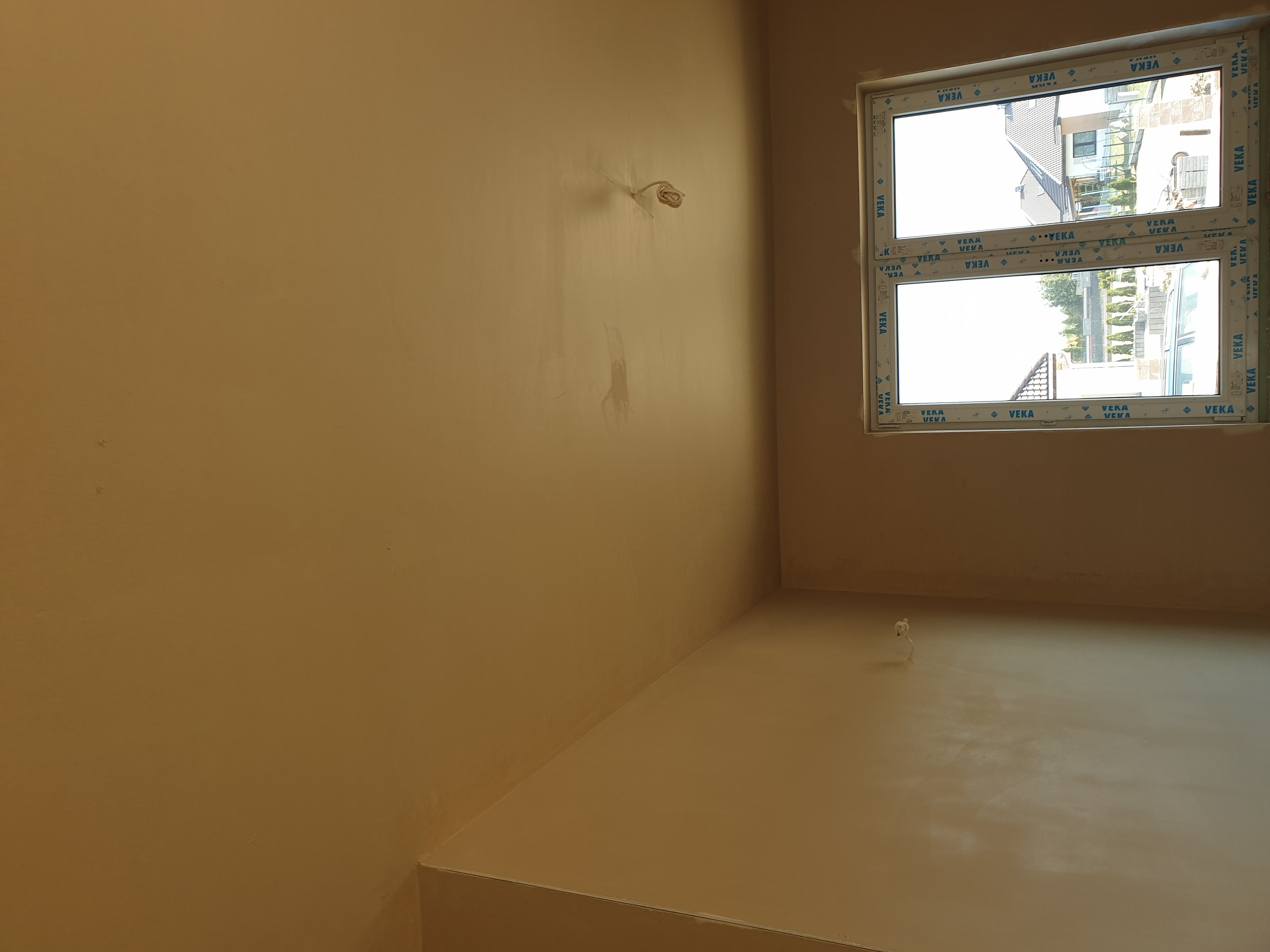 Świeżo położone, jednolite tynki na ścianach w pomieszczeniu z oknem PCV marki VEKA, widoczne wystające kable instalacji elektrycznej.