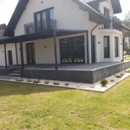 Nowoczesny dom jednorodzinny z białą elewacją, czarnymi ramami okiennymi i balkonem, otoczony zielonym trawnikiem i szarą kostką brukową.