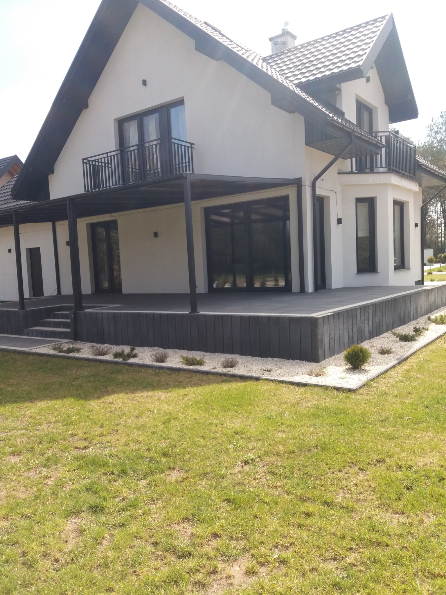 Nowoczesny dom jednorodzinny z białą elewacją, czarnymi ramami okiennymi i balkonem, otoczony zielonym trawnikiem i szarą kostką brukową.