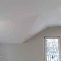 Poddasze po remoncie: gładkie, szare ściany i biały sufit ze skośnym fragmentem, okno z ozdobną balustradą i widokiem na ośnieżone drzewa, dwa punktowe reflektory sufitowe.