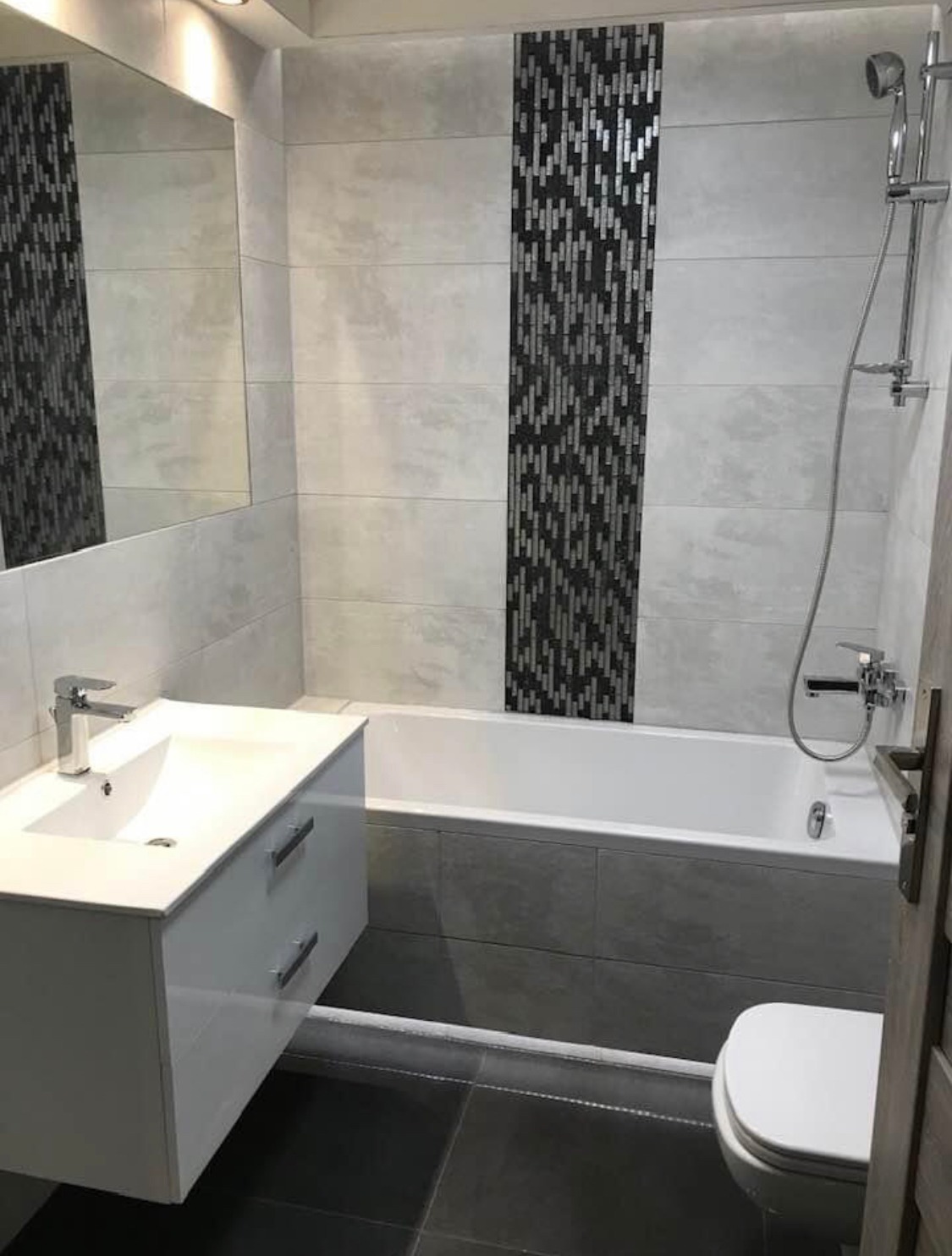 Wykończona łazienka z szarymi płytkami, dekoracyjnym pasem mozaiki, białą armaturą i podwieszaną szafką umywalkową.