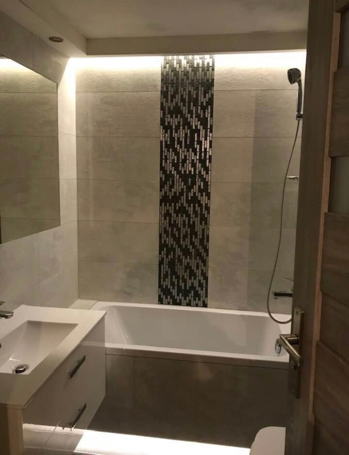 Nowoczesna łazienka z podświetleniem LED, beżowe płytki ścienne z dekoracyjnym pasem mozaiki, biała wanna i umywalka z szafką, widoczny fragment kabiny prysznicowej z deszczownicą.