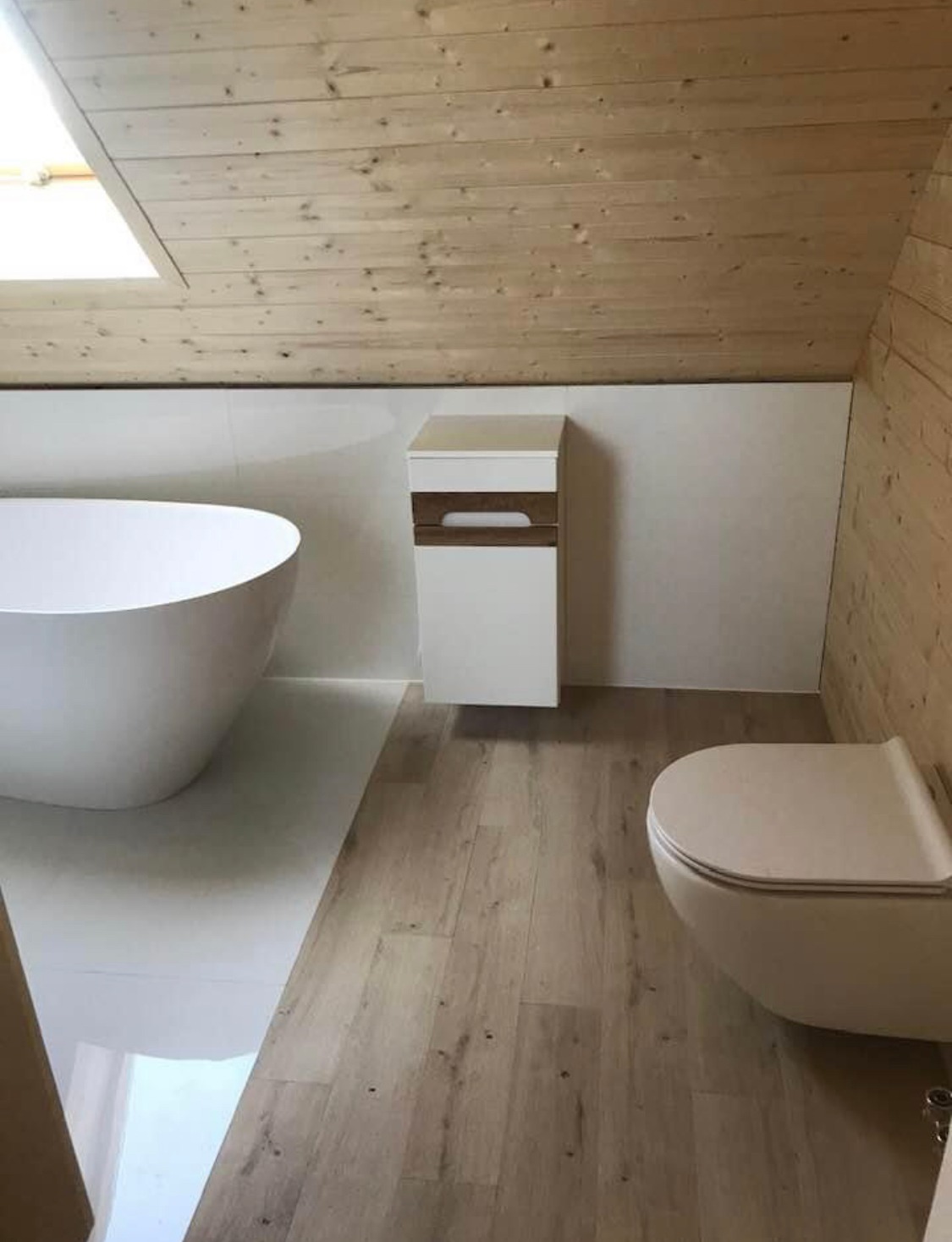 Nowoczesna łazienka na poddaszu z drewnianym wykończeniem sufitu i ściany, białą wanną wolnostojącą, podwieszaną toaletą i szafką umywalkową w minimalistycznym stylu, na podłodze panele imitujące...