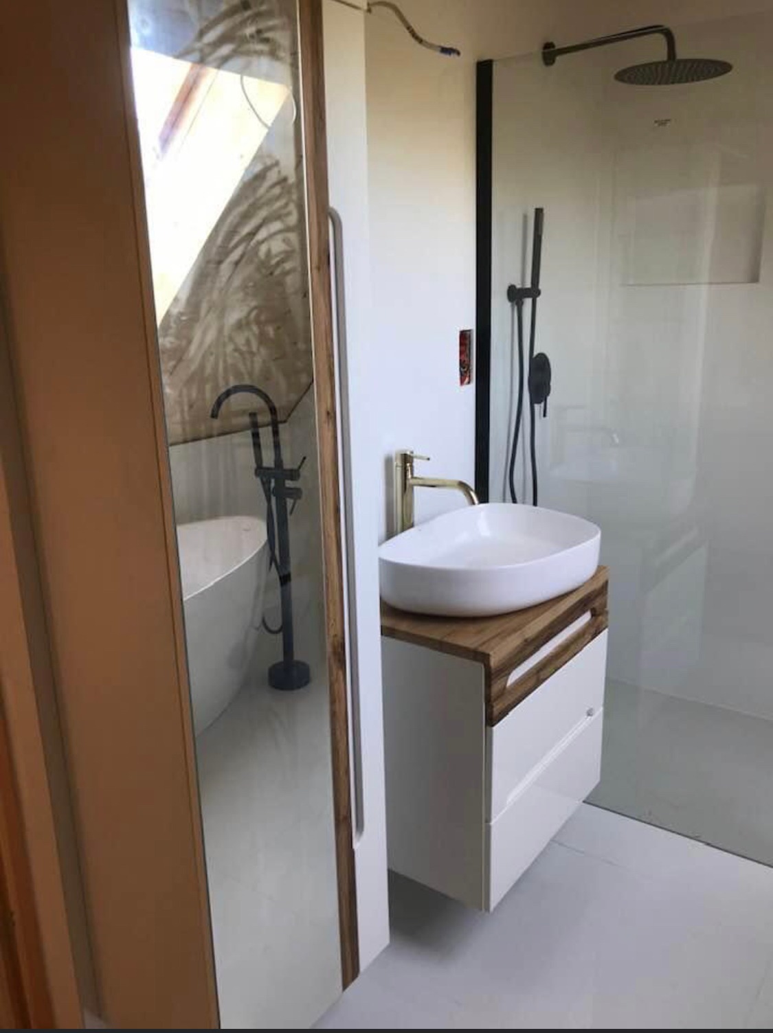 Nowoczesna łazienka z owalną umywalką nablatową, złotą baterią, kabiną prysznicową typu walk-in z czarną armaturą oraz odbiciem w lustrze wolnostojącej wanny z czarną baterią.