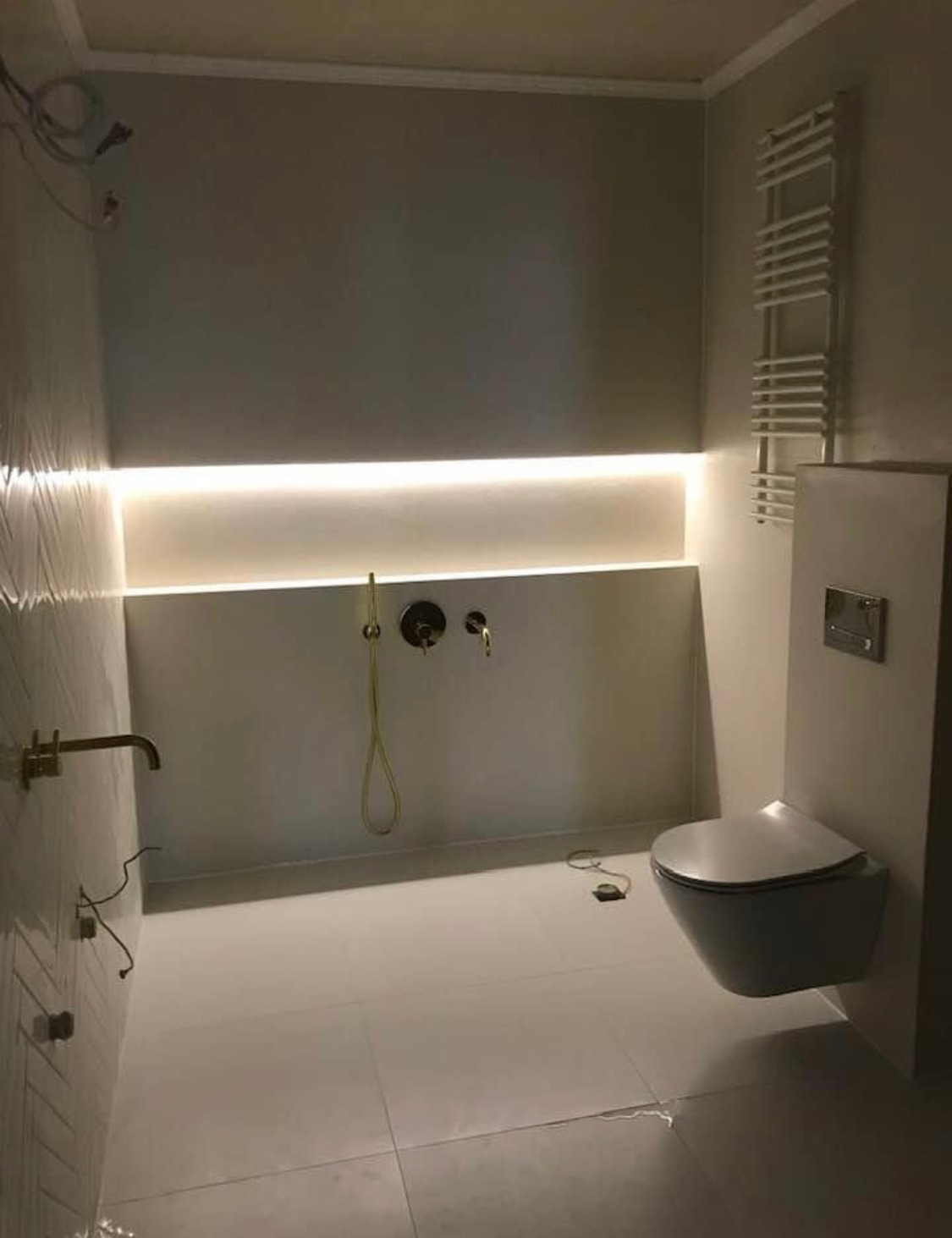 Nowoczesna łazienka w trakcie wykańczania, widoczne białe płytki podłogowe, podwieszana toaleta, grzejnik drabinkowy, złota armatura i oświetlenie LED w ścianie, z widocznymi kablami i elementami...