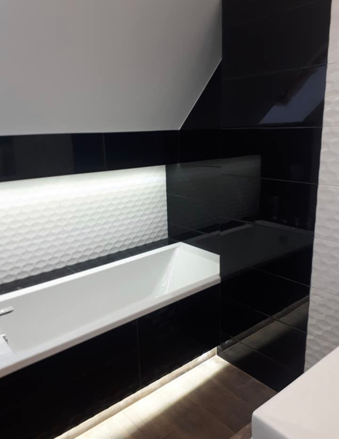 Nowoczesna łazienka z wanną, wykończona czarno-białymi płytkami o różnych fakturach, podświetlona listwą LED u dołu wanny.