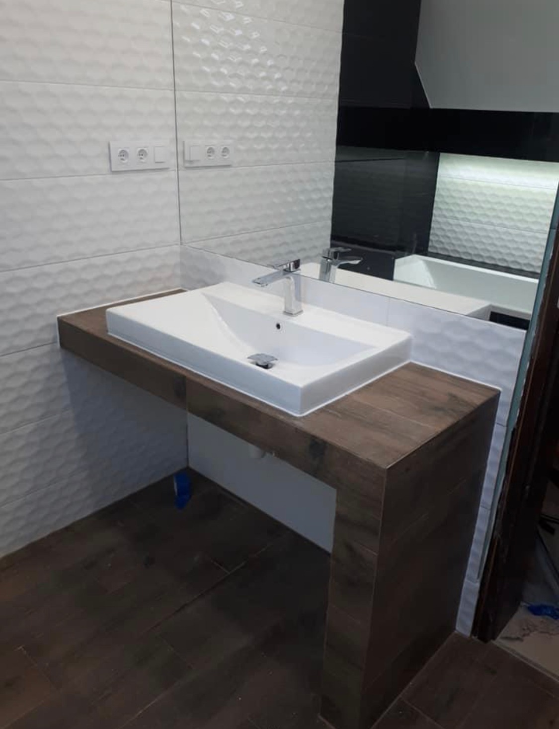 Nowoczesna łazienka z białą umywalką nablatową na blacie imitującym drewno, białe płytki strukturalne na ścianie i duże lustro.